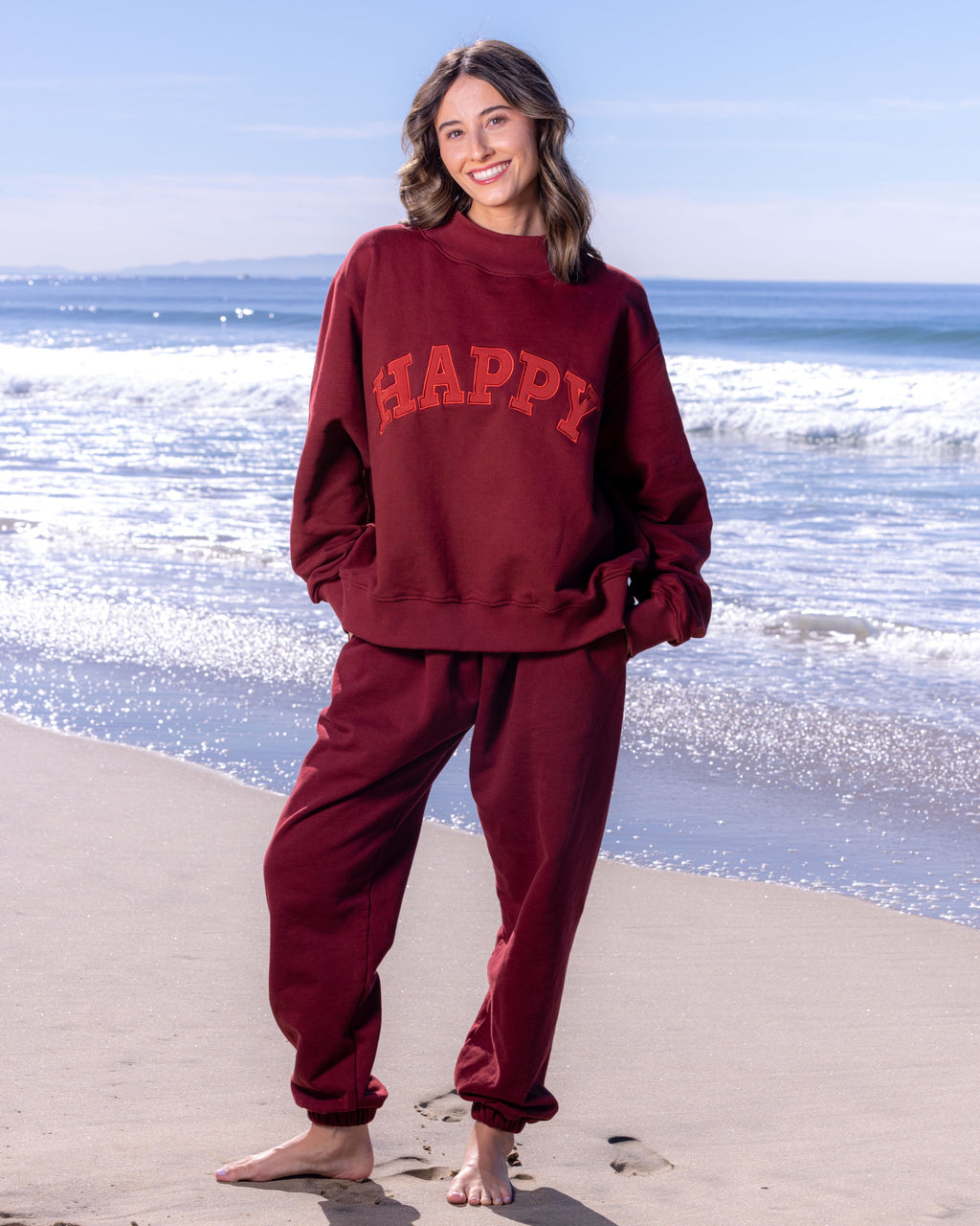 Person wearing a maroon 'HAPPY' sweatshirt and pants on a beach.