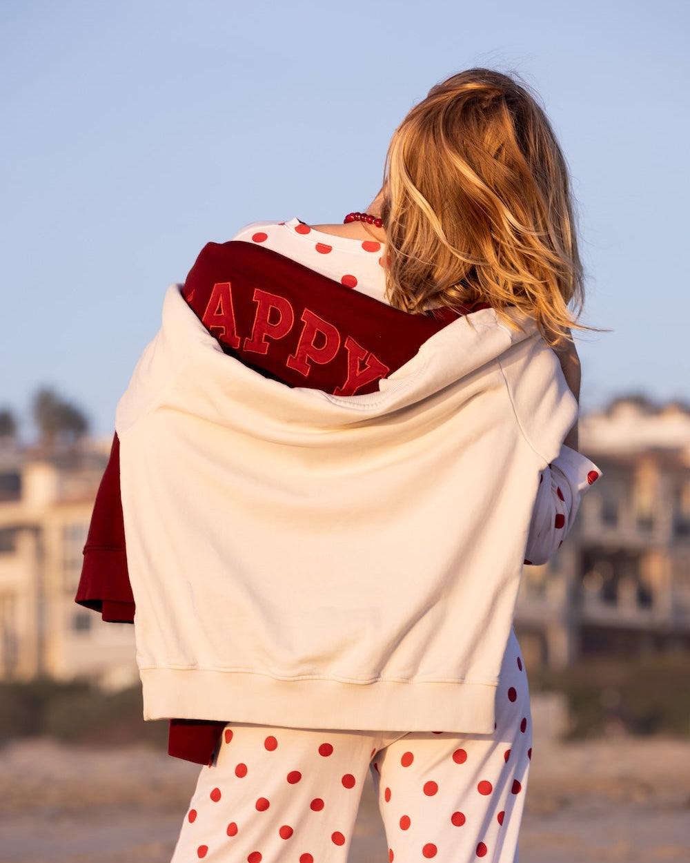Person wearing an off white sweatshirt with 'HAPPY' on the back, red mockneck sweatshirt, and red polka dot pants on a beach.