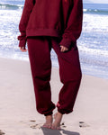 Woman in maroon 'HAPPY' sweatshirt and pants standing on a beach with ocean waves in the background.