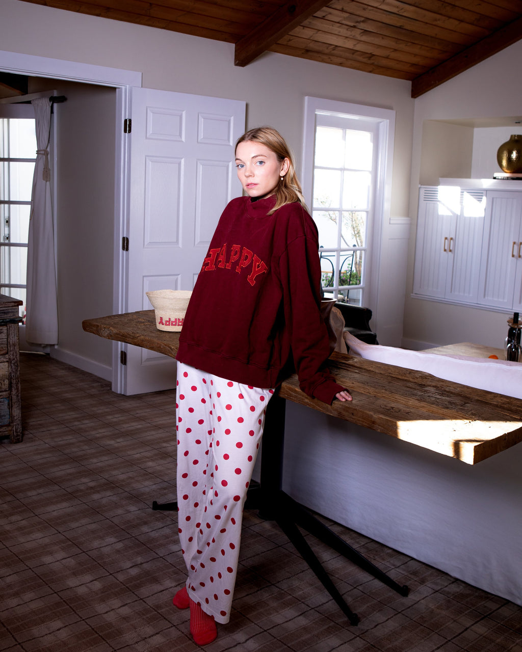 Woman in a HAPPY red sweatshirt and red polka dot pants standing in a room with wooden floors and white walls and a HAPPY straw hat on the table. 